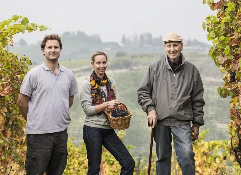 Three people standing among grapevines in Piedmont, one holding a basket of grapes